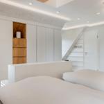 Schlafzimmer mit weißem Bett, Schrank und Treppe zur oberen Etage.
