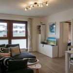 Wohnzimmer mit Sofa, Fernseher und Essbereich. Fenster mit Vorhängen und Holzboden.