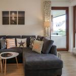 Ein Wohnzimmer mit grauem Sofa, Kissen und einem runden Tisch. An der Wand hängt ein Bild.