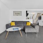 Wohnzimmer mit grauem Sofa, gelben Kissen und weißem Couchtisch. Küche und Treppe im Hintergrund.
