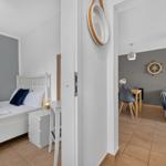 Schlafzimmer mit weißem Bett und Nachttisch. Durchgang in Wohnbereich mit Tisch, Stühlen und Sofa.