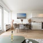 Dining area with table and chairs. Kitchen with cabinets and dishwasher. Large windows and wooden floor.