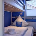 Ein Bett mit weißer Bettwäsche und blauem Kissen unter einem Doppelstockbett. An der Wand hängt eine Lampe.