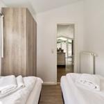 Zwei Betten mit weißen Laken und Handtüchern. Holzschrank und Fenster. Blick in die Küche.