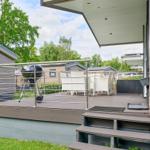 Terrasse mit Grill, weißem Gartenmöbel und Holzstufen. Hintergrund: Wohnwagen und Bäume.