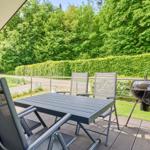 Terrasse mit Tisch, Stühlen und Grill. Hintergrund: grüne Hecke und Wald.