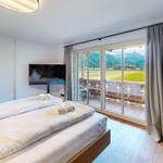 Schlafzimmer mit Balkonblick auf Berge und Grünflächen. Bett, TV und Wandbild.