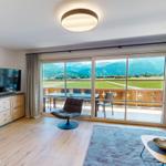 Wohnzimmer mit TV, Sitzgelegenheiten und Balkonblick auf grüne Landschaft und Berge.