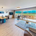 Wohn-Essbereich mit Balkon und Bergblick. Holzboden, Sofa, Esstisch und TV.