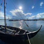 Ein schwarzes Segelboot liegt im Hafen. Im Hintergrund sind weitere Boote und ein Himmel mit Sonne und Wolken zu sehen.