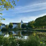 Schloss mit Turm spiegelt sich im Teich. Grüne Bäume und Sträucher umgeben das Wasser.