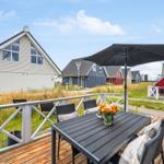Terrasse mit Tisch, Stühlen und Sonnenschirm. Hintergrund: mehrere Häuser und Grasland.