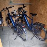 Zwei blaue E-Bikes stehen in einem Holzraum mit Steinboden und Stuhl.