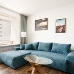 Ein Wohnzimmer mit blauem Sofa, Glas-Tisch und Fensterblick auf Gebäude.