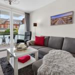 Wohnzimmer mit grauem Sofa, roten Kissen und Balkon mit Blick auf Gebäude.