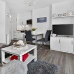 Wohnzimmer mit TV, Esstisch und Küche im Hintergrund. Weiße Möbel und Holzboden.
