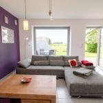 Wohnzimmer mit grauem Sofa, Holztisch und lila Wand. Durchgänge zu Terrasse und Garten.