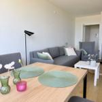 Ein Essbereich mit Holztisch und grauen Stühlen. Hinter dem Tisch ein grauer Sofa mit Kissen.
