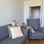 Ein Wohnbereich mit grauem Sofa, Sessel und Beistelltisch. Auf dem Sofa liegen Kissen.
