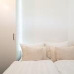 Doppelbett mit Kissen und Bettwäsche in einem hellen Raum mit Fenster und Schrank.