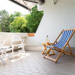 Terrasse mit zwei weißen Stühlen und einem blau-weißen Liegestuhl. Hintergrund: grüne Bäume.