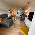 Wohnzimmer mit Sofa, Sessel und Küchenbereich. Holzboden und gelber Stuhl.