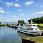 Ein weißes Schiff mit grüner Linie liegt am Hafen. Im Hintergrund sind Bäume und ein kleiner Leuchtturm zu sehen.
