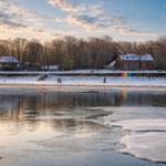 Schneebedeckter Uferbereich mit gefrorenem Wasser und rotem Gebäude im Hintergrund.