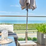Terrasse mit Blick auf das Meer und Grasland. Sitzmöbel, Sonnenschirm und Pflanzen.