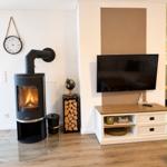 Wohnzimmer mit Kamin, Fernseher und Holzstapel. Wanduhr und Regal sichtbar.