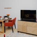 Ein Essbereich mit Holztisch, zwei orangefarbenen Stühlen und Fernseher auf einem Schrank.