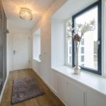 Hallway with wooden floor, mirror, window, and rug. Ceiling light and flowers on windowsill.