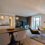 Dining area with table and chairs, living room with armchairs and TV, large windows with balcony view.
