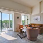 Living room with sofa, chairs, and balcony. Bedroom with bed in background.