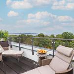 Terrasse mit Liege, Tisch und Obstschale. Blick auf Wald und See unter blauem Himmel.