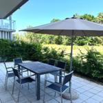 Terrace with table, four chairs, and umbrella. Background: grassy area and trees.