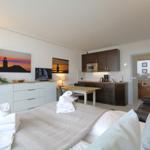 A bedroom with a bed, TV, and kitchen area in the background.
