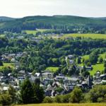 Panoramablick auf ein Dorf mit Häusern, Wiesen und bewaldeten Hügeln.