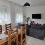 Wohn- und Essbereich mit Holztisch, Sofa und Fernseher. Fenster mit weißen Vorhängen.