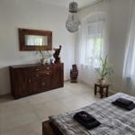 Schlafzimmer mit Bett, Holzschrank, Spiegel und Pflanzen. Fenster mit weißen Vorhängen.