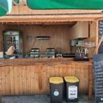 Holzbar mit Getränkeautomat und Preisliste. Zwei Mülltonnen stehen vor der Theke.