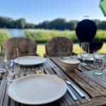Set table for four on wooden deck by lake. Grill and green umbrella visible.