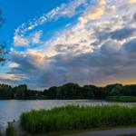 Lake with grassy strips and trees under a cloudy sky with moon.