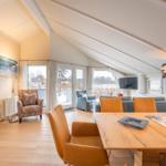 Living and dining area with wooden table, chairs, and TV. View of balcony and landscape.