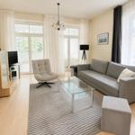 Wohnzimmer mit Sofa, Sessel, Glas-Tisch und großen Fenstern.