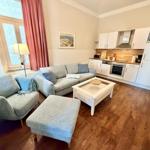Wohnzimmer mit Sofa, Sessel und Küchenbereich. Holzboden und Fenster mit roten Vorhängen.