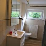 Badezimmer mit Waschbecken, Spiegelschrank, Toilette und Fenster mit Blick auf Grün.