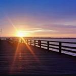 Wooden pier extends over water to sunset with warm light rays.
