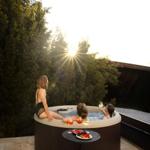 Drei Personen genießen einen Whirlpool auf einer Terrasse mit Blick auf Bäume und Sonnenuntergang.