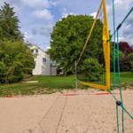 Sandplatz mit Volleyballnetz vor einem Haus im Grünen.